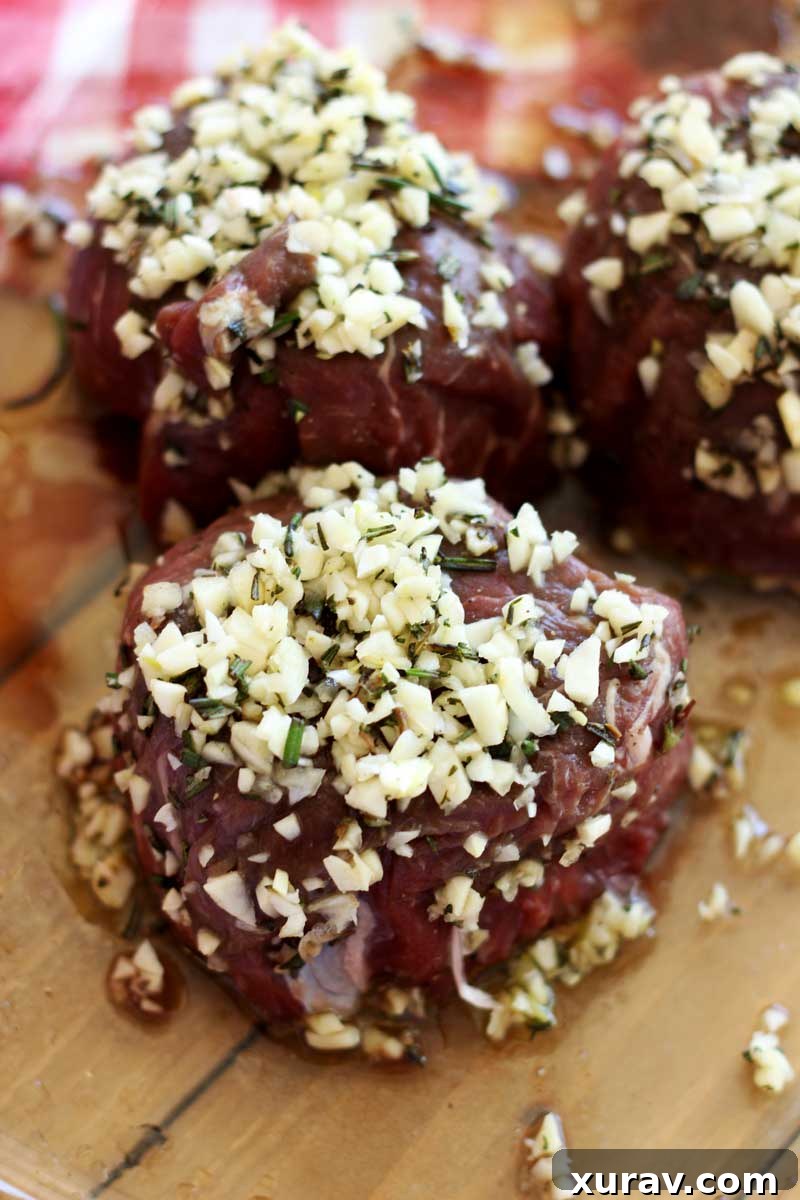 Close-up of a raw filet mignon steak covered generously with a fresh garlic and rosemary marinade in a glass dish.
