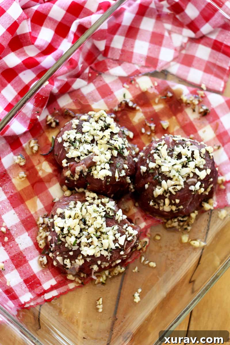 Marinated filet mignon steaks resting in a clear glass dish, showcasing the vibrant rosemary and minced garlic.
