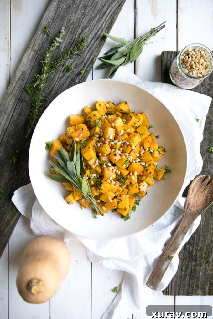 A large serving bowl overflowing with oven roasted garlic and herb butternut squash, garnished with fresh herbs and pine nuts