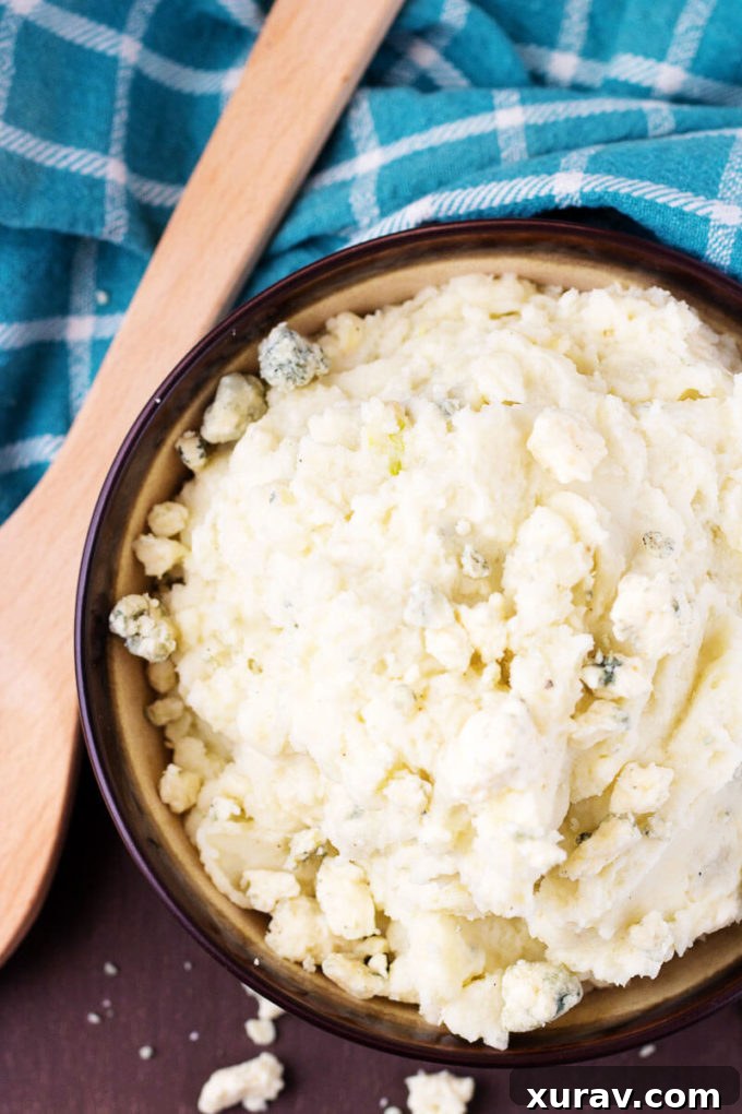 Top down view of a bowl of Roasted Garlic Bleu Cheese Mashed Potatoes with a wooden spoon, garnished and ready to serve.