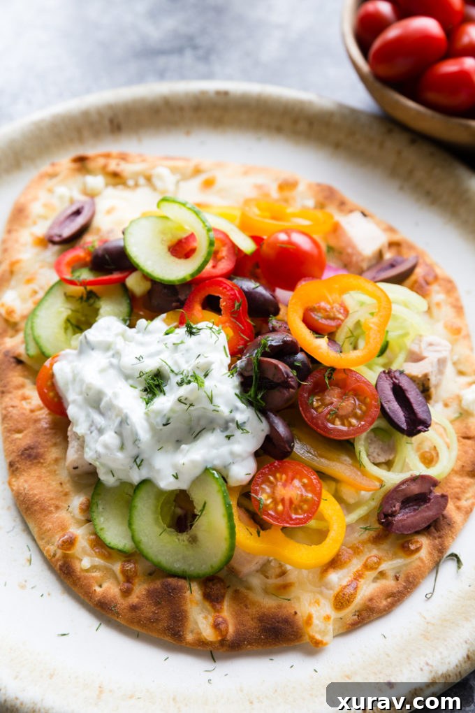 greek chicken flatbread on a cream colored plate. Topped with peppers, kalamata olives, cucumbers, tomatoes and cheese. With a sauce.