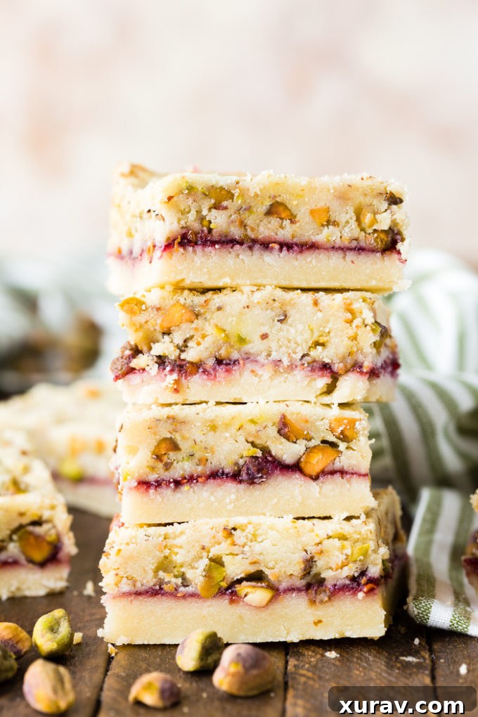 A stack of beautifully layered raspberry pistachio shortbread bars, showing the shortbread, jam, and pistachio layers.