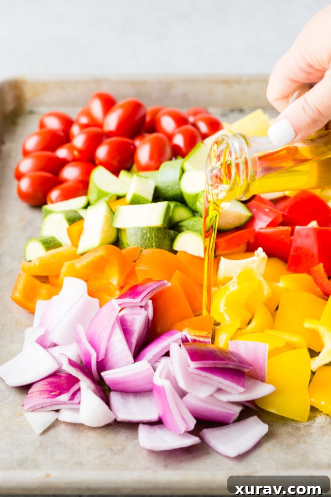Pouring olive oil over chopped Greek vegetables on a sheet pan, preparing them for roasting.