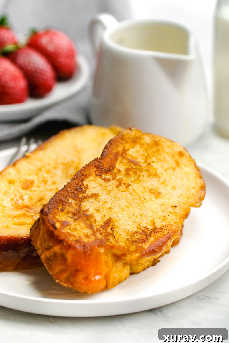 White plate with two slices of french toast, strawberries in the background, and a jug of syrup