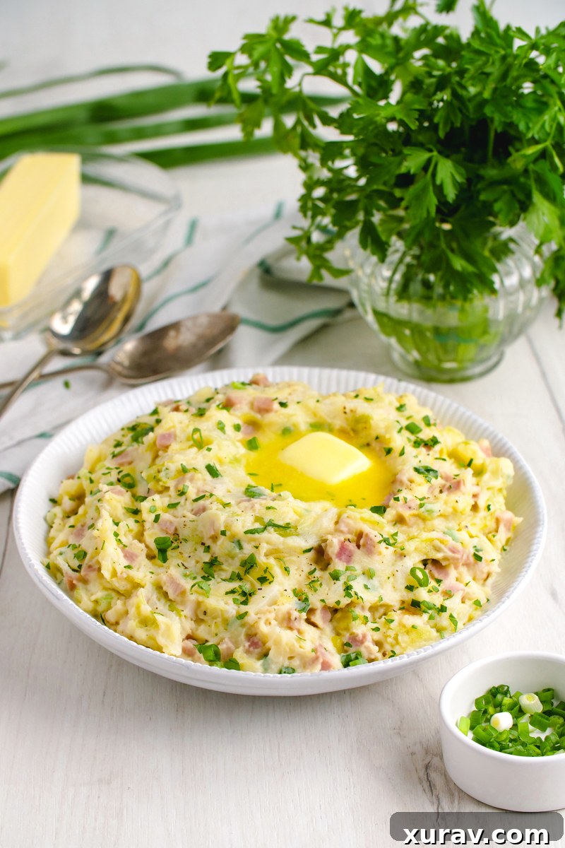 A white bowl filled with colcannon and topped with a pad of butter. Two spoons behind the colcannon and a vase of fresh parsley