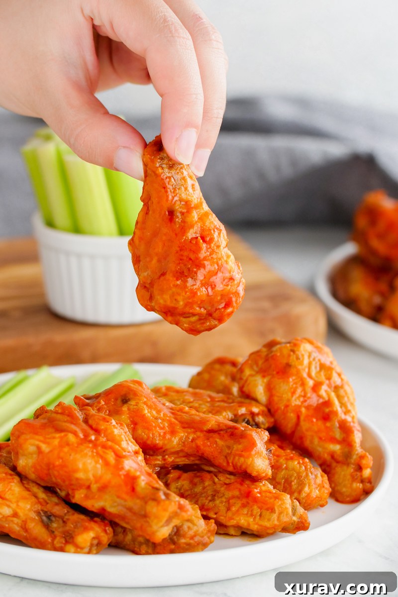 Buffalo wings, these hot wings are on a white plate with celery in the background, and a hand holding one chicken wing up.