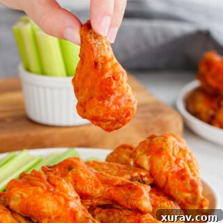 Buffalo wings, these hot wings are on a white plate with celery in the background, and a hand holding one chicken wing up.