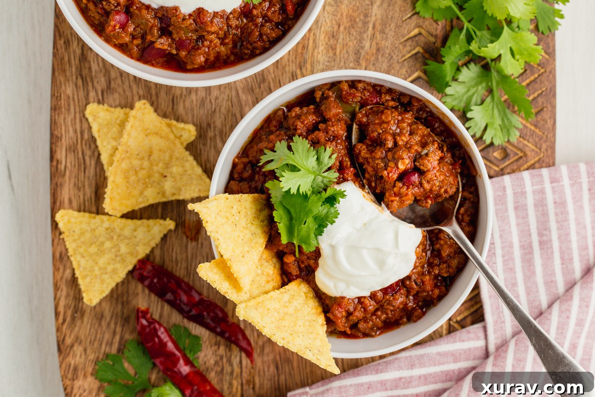 A generous serving of Chili Con Carne in a white bowl, topped with shredded cheese, a dollop of sour cream, and fresh green onions, against a rustic backdrop.