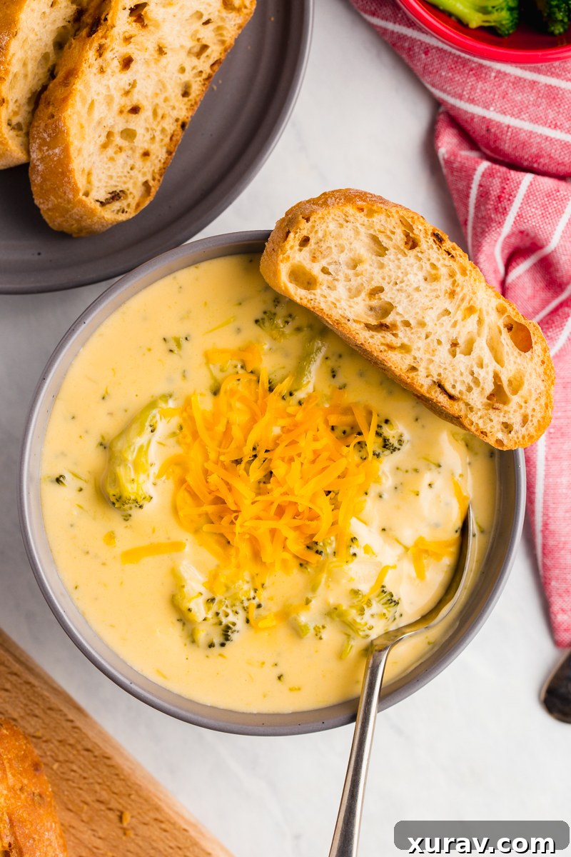 A bowl of creamy broccoli cheese soup garnished with fresh parsley and shredded cheddar cheese, with a spoon dipping in.