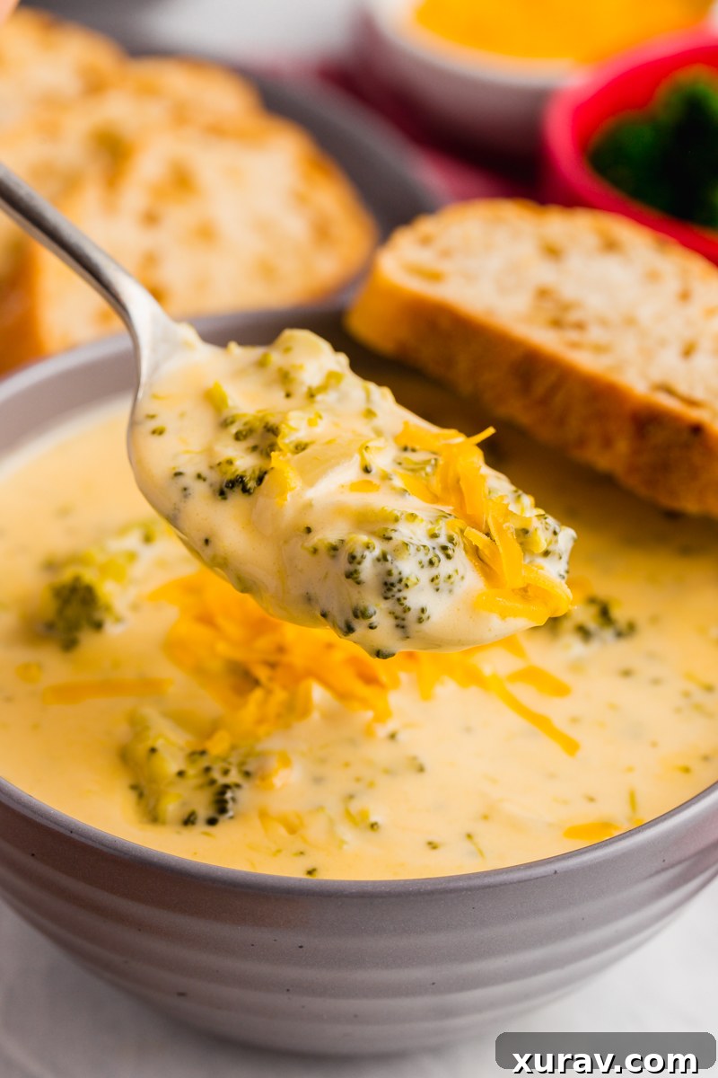 A bowl of creamy broccoli cheese soup topped with a swirl of cream or cheese, with a rustic breadstick on the side.