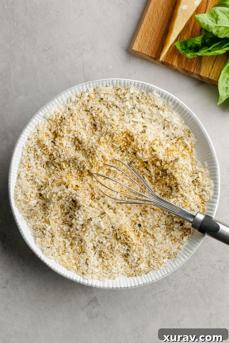 Two hands dipping a raw chicken breast into a bowl of seasoned bread crumbs.