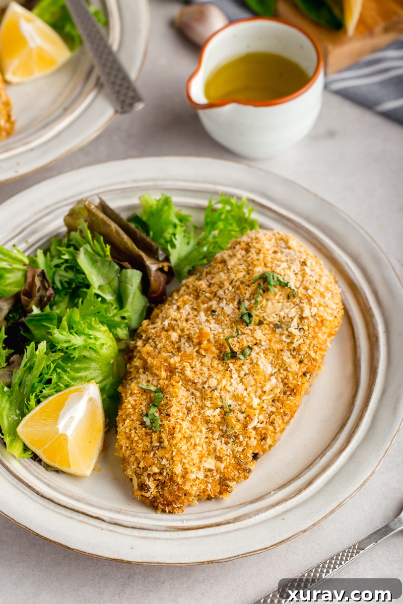 Six baked breaded chicken breasts stacked on a serving plate with fresh herbs and a lemon wedge.