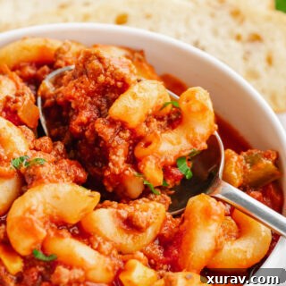 A bowl of American Goulash, white bowl, silver spoon, elbow macaroni, and some green garnish on top. Bread in the background.