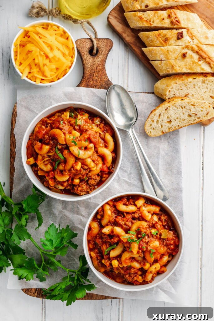 Two generous bowls of Instant Pot American Goulash, garnished with fresh herbs and served with a side of crusty bread, cheese, and sour cream.