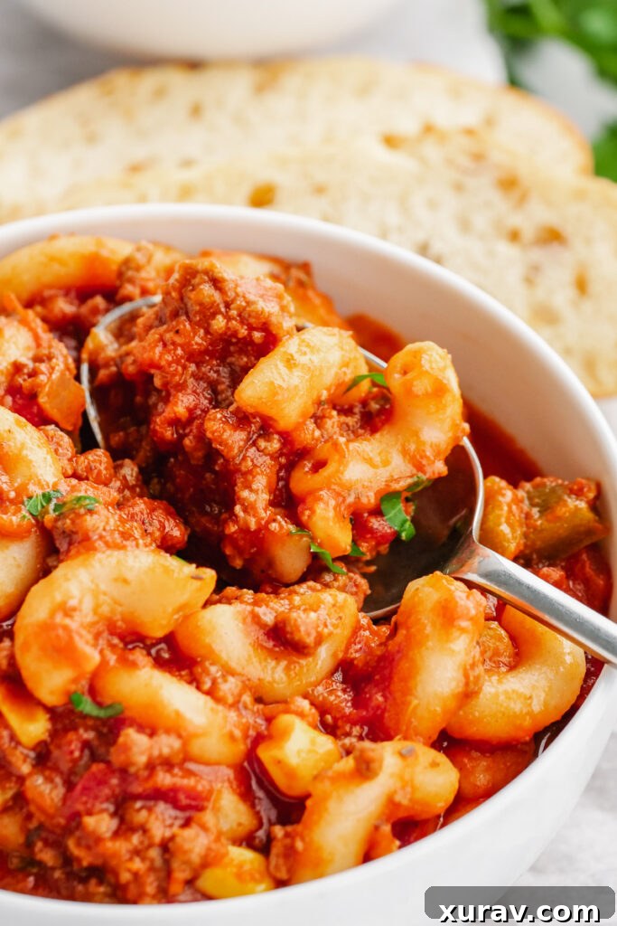 A pristine white bowl filled with perfectly prepared Instant Pot American Goulash, showcasing tender elbow macaroni, savory beef, and a rich tomato sauce, garnished with fresh herbs. A side of crusty bread is blurred in the background.
