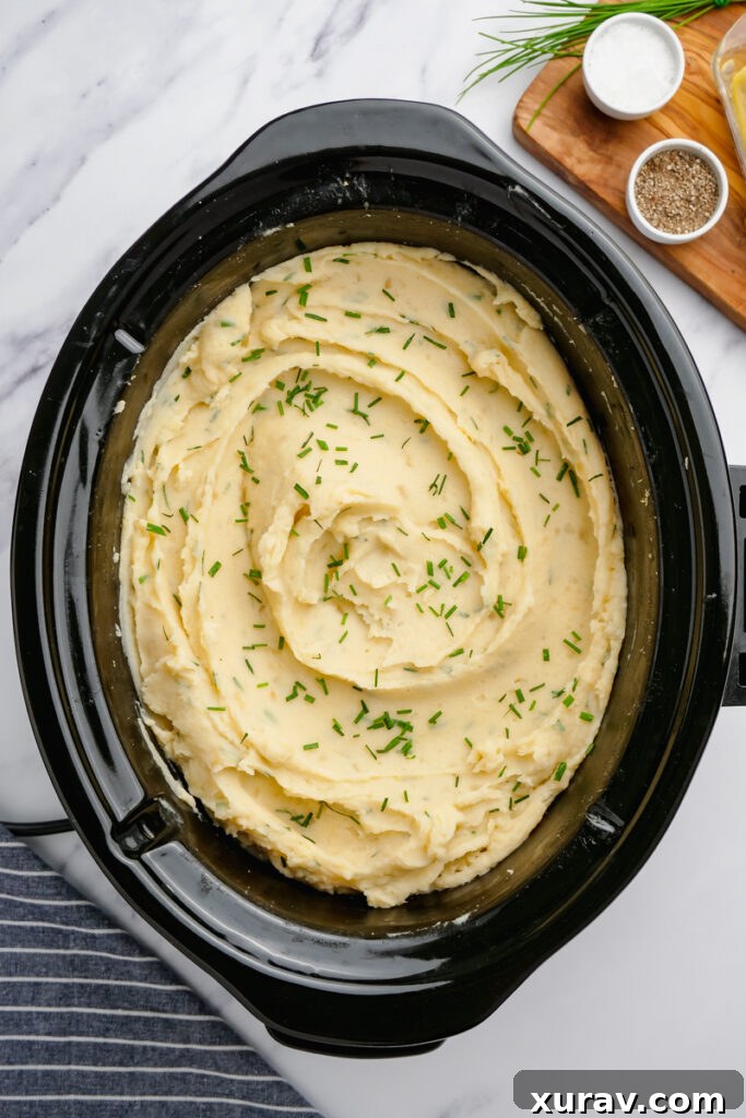 Slow Cooker Mashed Potatoes, ready to serve in a black slow cooker, garnished with fresh chives.