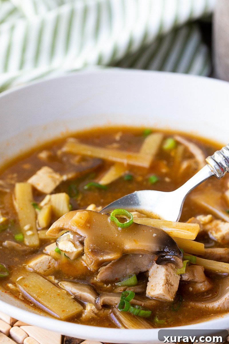 A steaming bowl of hot and sour soup filled with tender tofu cubes, thinly sliced pork, bamboo shoots, and mushrooms in a rich, golden-brown broth. Swirls of silky egg ribbons float on top, garnished with fresh scallions and a glisten of chili oil for a spicy kick. Served in a white ceramic bowl, the soup looks hearty, comforting, and full of bold flavors.