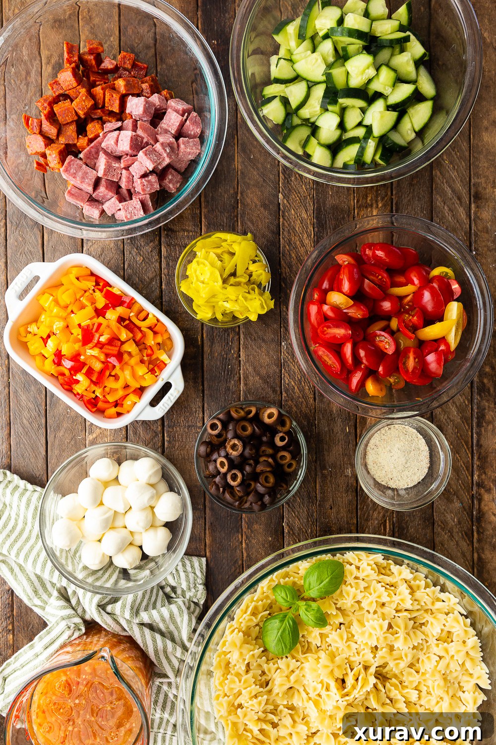A beautiful array of fresh ingredients laid out, including pasta, pepperoni, salami, mozzarella, olives, tomatoes, cucumbers, and peppers, ready for an easy pasta salad.